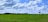 A vast green agricultural field stretches towards the horizon under a bright blue sky with scattered white clouds. Rows of lush plants cover the ground, suggesting cultivated crops. A few solitary trees can be seen, and low hills are visible in the distance.