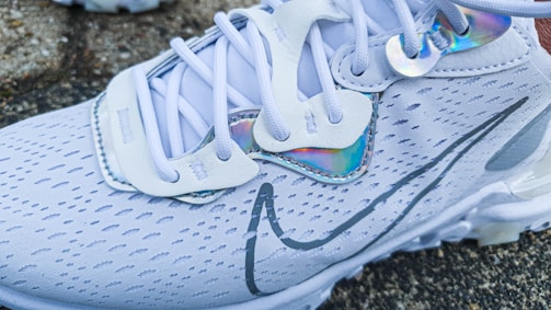 A close-up of a stylish white athletic shoe with intricate patterns and shoelaces. The shoe features a sleek design with iridescent accents near the eyelets and on a prominent logo.