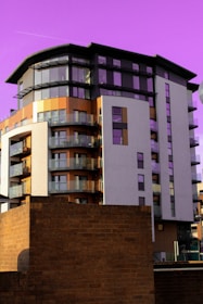 A modern, multi-story apartment building with a combination of glass balconies, beige, and brown exterior finishes stands prominently against a vivid purple sky. In the foreground, a brick structure partially obstructs the view of the building's base.