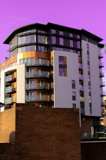 A modern, multi-story apartment building with a combination of glass balconies, beige, and brown exterior finishes stands prominently against a vivid purple sky. In the foreground, a brick structure partially obstructs the view of the building's base.