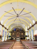 An ornate church interior with wooden pews leading to an intricately carved altar. The ceiling is arching with a large ceiling fan at the center. Stained glass windows align the walls, and religious statues and decorative pieces add to the sacred atmosphere.