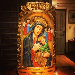 A colorful religious icon depicting a crowned woman holding a child, surrounded by angels. The image features intricate details with vibrant colors, set in a rustic wooden frame with ornate carvings.