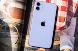 A smartphone with a prominent apple logo on its back is placed on top of an open magazine. The smartphone is white with dual cameras. The background includes a photograph of a blue bicycle with yellow wheel rims and a person holding it, along with a red and white checkered fabric bag.