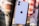 A smartphone with a prominent apple logo on its back is placed on top of an open magazine. The smartphone is white with dual cameras. The background includes a photograph of a blue bicycle with yellow wheel rims and a person holding it, along with a red and white checkered fabric bag.
