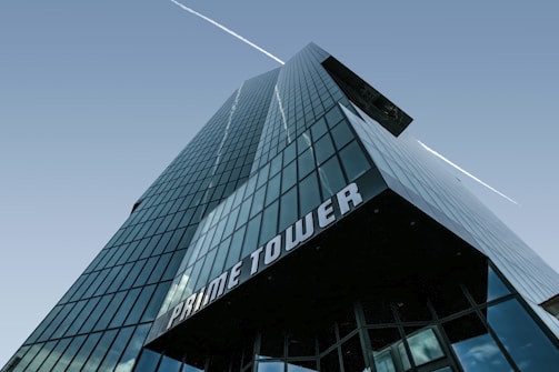 A modern skyscraper with reflective glass panels, towering into the clear sky. The building has a sleek, angular design with the words 'PRIME TOWER' prominently displayed near its base. A white contrail is visible in the sky, running parallel to the building.
