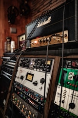 A close-up view of audio equipment with various control panels, switches, and knobs. A prominent guitar amplifier with a recognizable logo is stacked above a series of audio processing units. The setup suggests a professional music production or recording studio environment. Coiled cables are visible, connecting the devices.