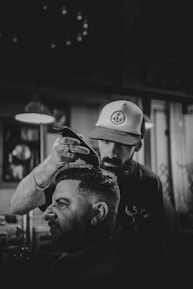 A barber is cutting a man's hair in a dimly lit barbershop, using electric clippers. The barber wears a hat with an anchor design and concentrates on his work. The atmosphere is casual and focused, with several blurred light fixtures and framed items visible in the background.