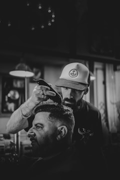 A barber is cutting a man's hair in a dimly lit barbershop, using electric clippers. The barber wears a hat with an anchor design and concentrates on his work. The atmosphere is casual and focused, with several blurred light fixtures and framed items visible in the background.