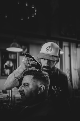 A barber is cutting a man's hair in a dimly lit barbershop, using electric clippers. The barber wears a hat with an anchor design and concentrates on his work. The atmosphere is casual and focused, with several blurred light fixtures and framed items visible in the background.