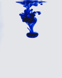 Vibrant blue ink swirls gently through a clear liquid, creating delicate, abstract shapes against a soft, light background. The motion captures the fluid dynamics as the ink disperses gradually.
