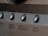 A close-up view of a brushed metal panel featuring several rounded buttons. Each button has a label, including 'ZONE 2/3/REC SELECT' and 'VIDEO SELECT'. The panel has an industrial and polished appearance with a smooth texture.