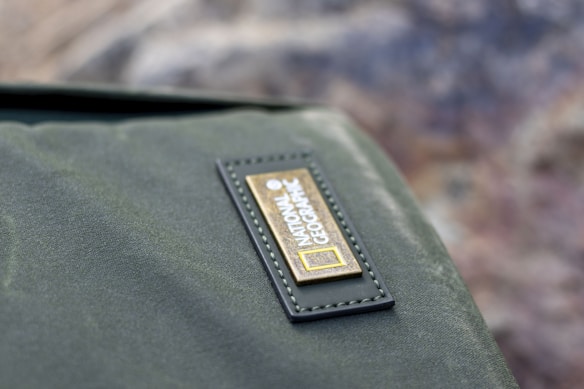 A close-up view of a green fabric item with a National Geographic logo on a small rectangular leather patch. The patch is sewn onto the fabric, and the background appears blurred with earthy tones.