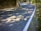 A winding road curves through a rural landscape, bordered by trees and grass. Sunlight filters through the leaves, casting dappled shadows on the road. White road markings indicate the lane edges, and there are reflective markers along the roadside. The road is well-paved and clear of debris.