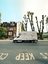 A white delivery van with graffiti covering its side is parked on a street lined with residential buildings and pruned trees. The pavement has clear road markings saying 'KEEP CLEAR'. The sky is clear and blue, with some buildings and green foliage visible in the background.