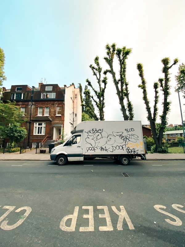 A white delivery van with graffiti covering its side is parked on a street lined with residential buildings and pruned trees. The pavement has clear road markings saying 'KEEP CLEAR'. The sky is clear and blue, with some buildings and green foliage visible in the background.