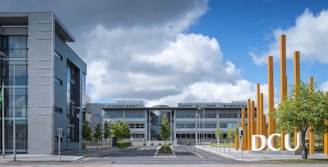Modern campus environment featuring a large building with a glass facade on the left and another multi-story building directly ahead. Stylized metal poles in an artistic arrangement stand to the right, with trees lining the road. The sky is partly cloudy, adding a dynamic element to the scene.