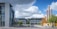Modern campus environment featuring a large building with a glass facade on the left and another multi-story building directly ahead. Stylized metal poles in an artistic arrangement stand to the right, with trees lining the road. The sky is partly cloudy, adding a dynamic element to the scene.