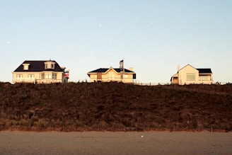 Three house-like buildings sit atop a small hill with a sandy ground in the foreground. The buildings are spaced out evenly with clear skies above.