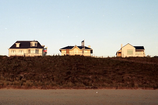 Three house-like buildings sit atop a small hill with a sandy ground in the foreground. The buildings are spaced out evenly with clear skies above.