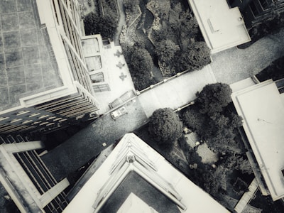 Aerial view of an urban area featuring several buildings and a pathway surrounded by greenery. It's a monochrome image with a large building on the left side, a smaller building on the right, and a pathway leading through landscaped gardens with trees and shrubs. A car is parked on the path, and various outdoor seating arrangements are visible.