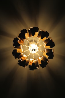 A ceiling light fixture emits rays of light, creating a starburst pattern against a dark background. The light is surrounded by a circular arrangement of metallic elements, which reflect the light and add an artistic visual interest.