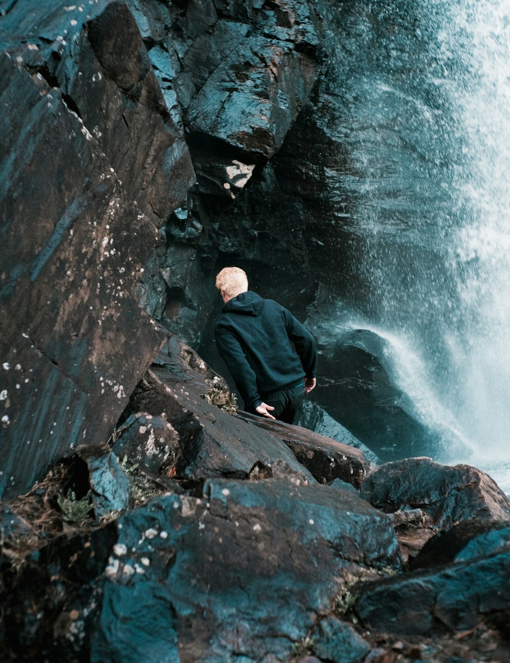 uomo in giacca nera che si siede sulla roccia vicino alle cadute d'acqua durante il giorno
