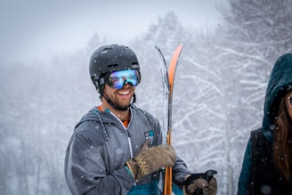 A person wearing a black helmet and reflective ski goggles holds a pair of skis while standing in a snowy forest. Snow is visibly falling, and the person seems cheerful with a broad smile. Another person with long hair and a dark hooded jacket is partially visible next to them.