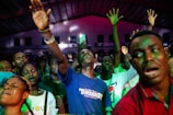 A group of people appears to be deeply engaged in an emotional experience, possibly during a concert or religious gathering. Many have their eyes closed and hands raised, suggesting a sense of euphoria or spiritual connection. The lighting is dynamic, casting colorful hues across the crowd.