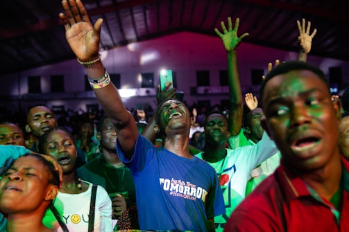 A group of people appears to be deeply engaged in an emotional experience, possibly during a concert or religious gathering. Many have their eyes closed and hands raised, suggesting a sense of euphoria or spiritual connection. The lighting is dynamic, casting colorful hues across the crowd.