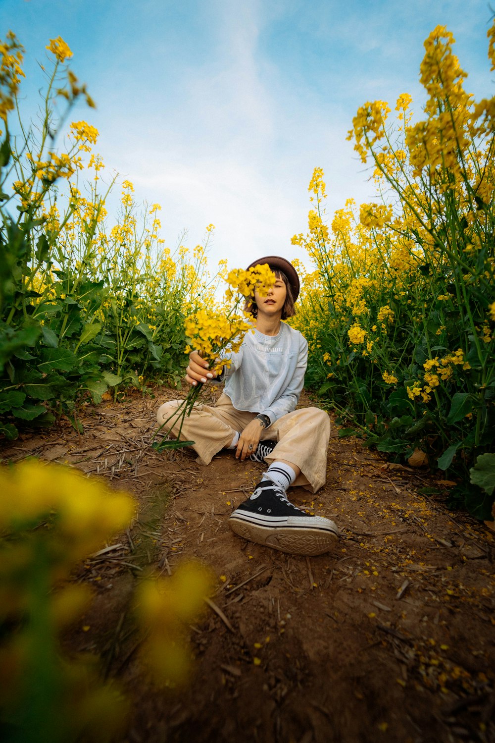 woman in white long sleeve shirt sitting on brown dirt road between yellow flowers during daytime