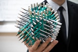 A person holds an intricate geometric structure made of numerous green pencils, held together with rubber bands. The person is dressed in a formal dark suit and tie, and the background is slightly blurred, suggesting an indoor setting.