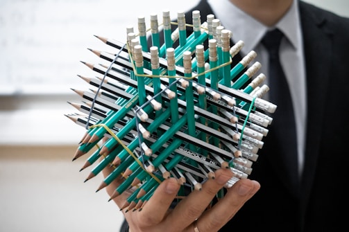 A person holds an intricate geometric structure made of numerous green pencils, held together with rubber bands. The person is dressed in a formal dark suit and tie, and the background is slightly blurred, suggesting an indoor setting.