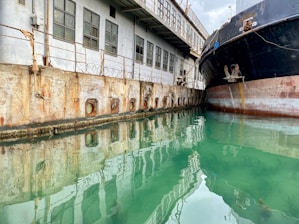 A large, slightly rusted ship is docked along a weathered industrial building with multiple windows and visible wire fencing above a concrete wall. The calm, greenish water reflects both the ship and the building.