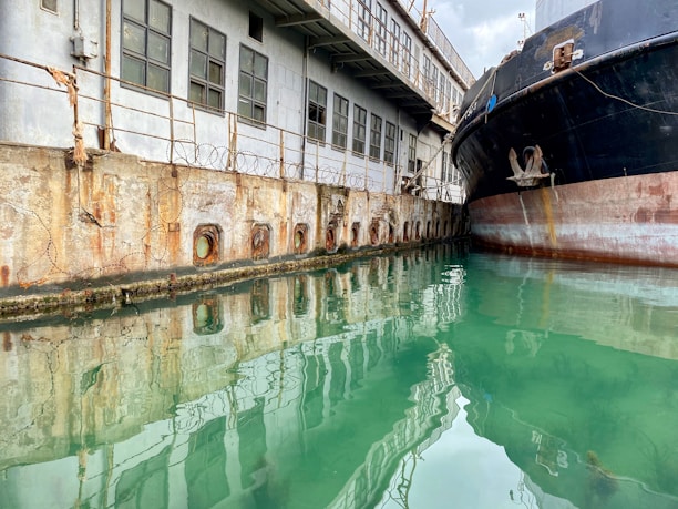 A large, slightly rusted ship is docked along a weathered industrial building with multiple windows and visible wire fencing above a concrete wall. The calm, greenish water reflects both the ship and the building.