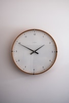 A simple, modern round wall clock with a wooden frame and a white face, featuring black hour and minute hands. The clock has a minimalist design with Arabic numerals marking the hours and subtle minute markers around the edge. It is set against a plain light-colored wall.