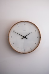A simple, modern round wall clock with a wooden frame and a white face, featuring black hour and minute hands. The clock has a minimalist design with Arabic numerals marking the hours and subtle minute markers around the edge. It is set against a plain light-colored wall.