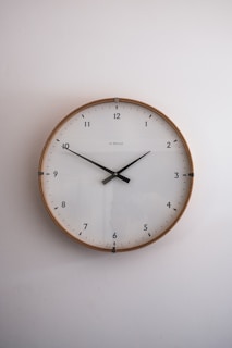 A simple, modern round wall clock with a wooden frame and a white face, featuring black hour and minute hands. The clock has a minimalist design with Arabic numerals marking the hours and subtle minute markers around the edge. It is set against a plain light-colored wall.