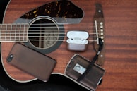 A wooden acoustic guitar lies flat with a smartphone, a pair of wireless earbuds in their case, and a leather wallet placed on its body. The items are arranged artfully, emphasizing the guitar's strings and wood grain texture.