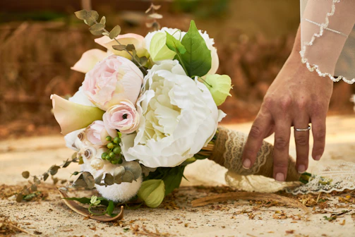 A bride's hand gently holding a vibrant bouquet of mixed roses in white, baby pink, and warm hues, radiating a soft, romantic glow.