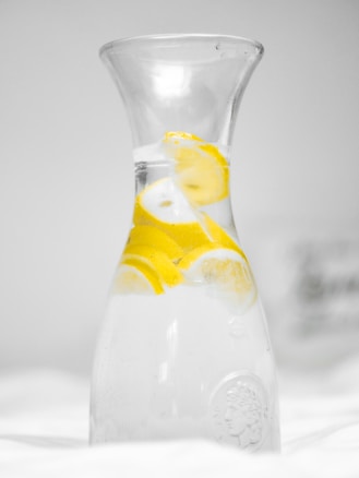 A clear glass carafe filled with water and several slices of lemon. The background is softly blurred and the overall color palette is light and minimalistic.