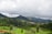 A lush green landscape filled with terraced plantations, surrounded by majestic hills partially veiled by low-hanging clouds. A small village with colorful houses lies nestled within the scenery, adding a touch of human presence to the natural setting.