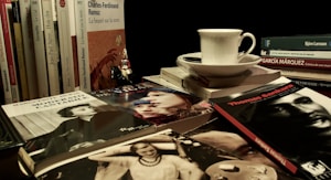 A collection of books is stacked horizontally and vertically with titles visible, featuring a variety of authors. A white coffee cup sits on top of one stack, accompanied by a saucer. A small figurine is placed among the books.