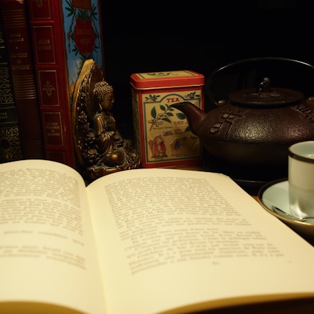An open book lies on a table, surrounded by decorative items. There is a small Buddha statue next to a collection of vintage red and brown books. A colorful tea tin and a traditional iron teapot add to the cozy ambiance. A white teacup with a saucer is in the foreground.