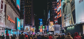 A bustling city street at night, crowded with people walking and surrounded by brightly lit billboards and advertisements. The vibrant lights illuminate various advertisements and company logos, creating a lively and dynamic atmosphere.