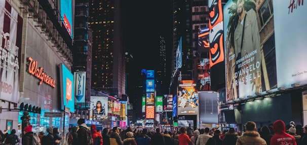 A bustling city street at night, crowded with people walking and surrounded by brightly lit billboards and advertisements. The vibrant lights illuminate various advertisements and company logos, creating a lively and dynamic atmosphere.