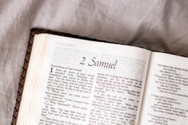 A close-up view of an open book showing a page from the Bible, specifically from the book of 2 Samuel. The text on the left page begins with a stylized chapter heading and includes verses that are clear and legible. The book rests on a soft, crumpled fabric surface, indicating a casual or relaxed setting.