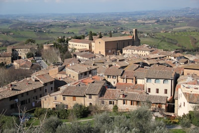A picturesque Italian hilltop town with charming, rustic buildings and terracotta roofs. A large historic church or monastery dominates the view, surrounded by lush green fields extending into the distance. The landscape has a serene and timeless quality, with rolling hills and scattered trees.