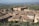 A picturesque Italian hilltop town with charming, rustic buildings and terracotta roofs. A large historic church or monastery dominates the view, surrounded by lush green fields extending into the distance. The landscape has a serene and timeless quality, with rolling hills and scattered trees.