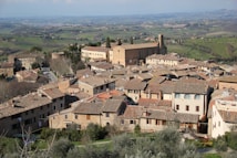 A picturesque Italian hilltop town with charming, rustic buildings and terracotta roofs. A large historic church or monastery dominates the view, surrounded by lush green fields extending into the distance. The landscape has a serene and timeless quality, with rolling hills and scattered trees.