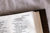An open book displaying text from Psalms 34:19, with a focus on the verse and surrounding text. The book's cover is partially visible, and there is soft lighting, suggesting a calm or introspective setting.